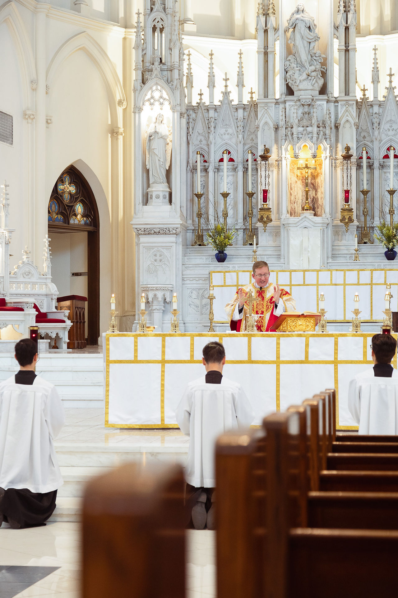 A priest in red robes holds mass at an ornate white altar. Three altar servers kneel in front of him in a church with carved details.