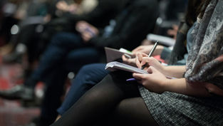 People seated in rows, taking notes on notepads with pens. Focus on hands and writing, suggesting a conference or lecture setting.