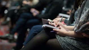 People seated in rows, taking notes on notepads with pens. Focus on hands and writing, suggesting a conference or lecture setting.