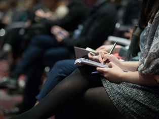 People seated in rows, taking notes on notepads with pens. Focus on hands and writing, suggesting a conference or lecture setting.