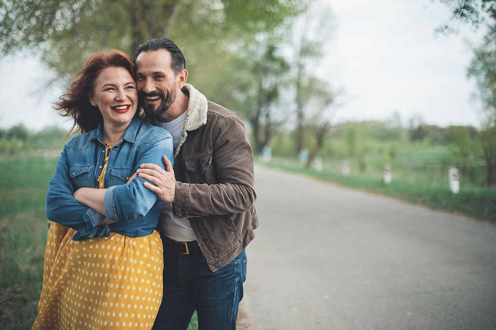 A smiling couple embraces on a rural road, surrounded by greenery. The woman wears a yellow polka dot dress, and the man wears a brown jacket.