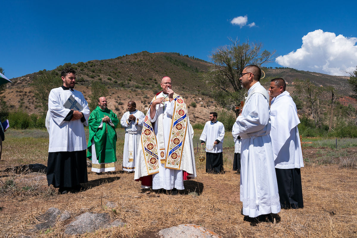 Priests in white and green vestments stand outdoors on a dry hillside under a blue sky, holding prayer items in a solemn ceremony