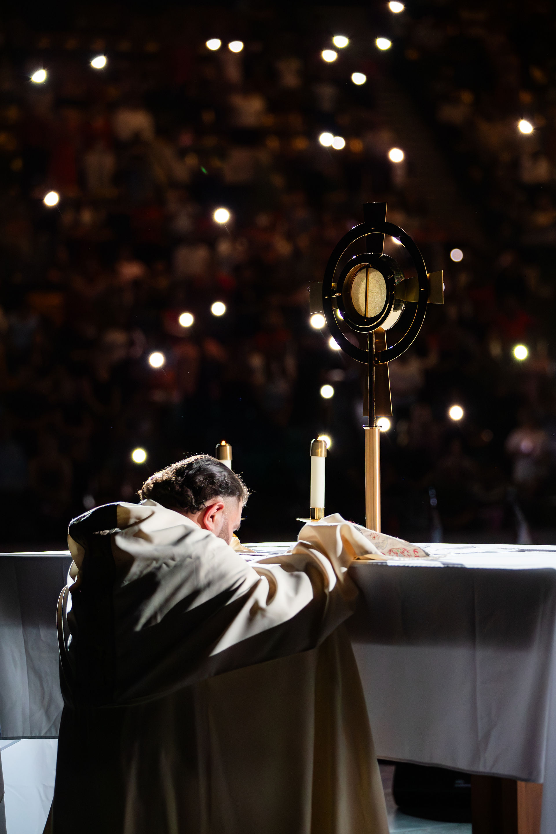 A priest kneels in prayer before the Eucharist on an altar, surrounded by a crowd holding lit candles in a dark setting.