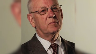 Portrait of an elderly man in a suit and glasses, looking serious against a plain beige background.