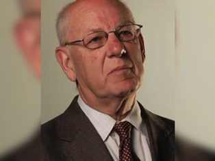 Portrait of an elderly man in a suit and glasses, looking serious against a plain beige background.