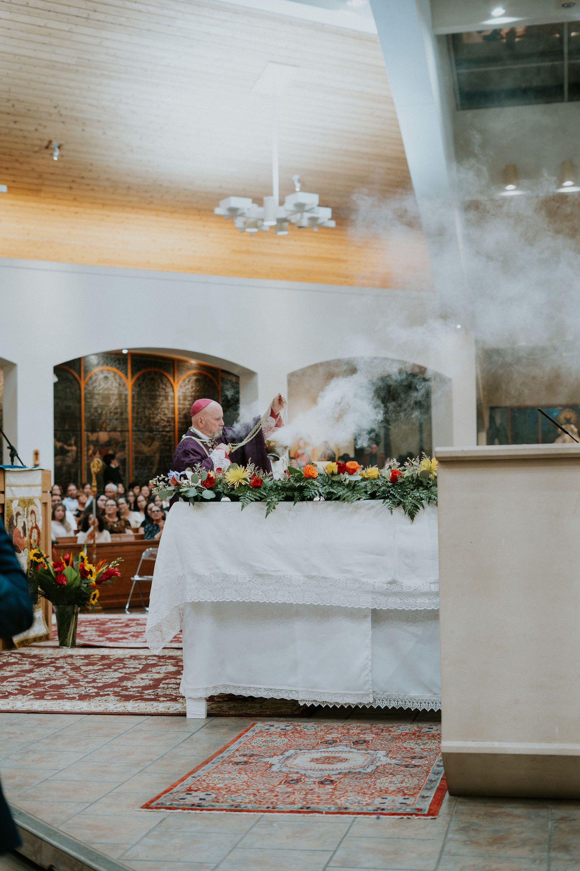 A clergyman in a purple robe swings an incense burner at an altar adorned with colorful flowers in a church. Congregation watches solemnly.