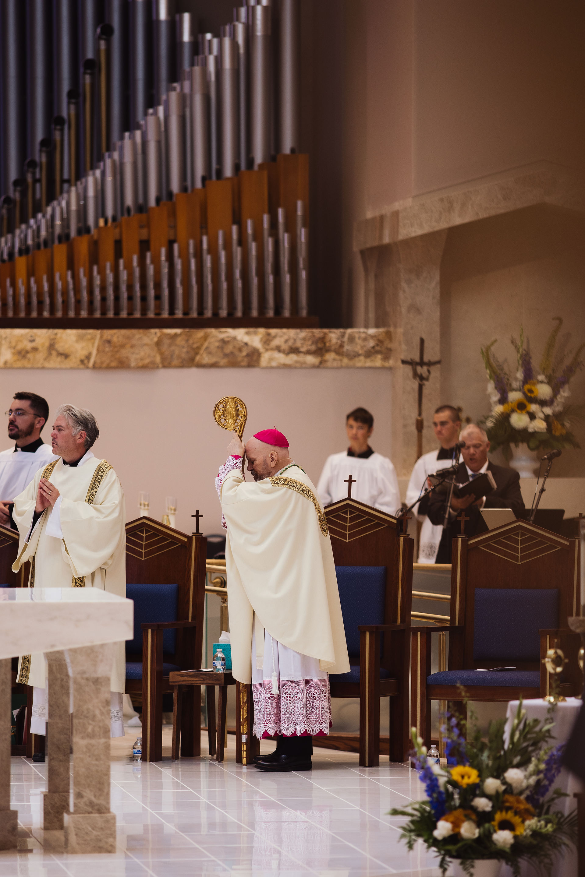 A bishop in a red cap holds a staff, surrounded by clergy in white robes during a church service. Background features organ pipes and flowers.