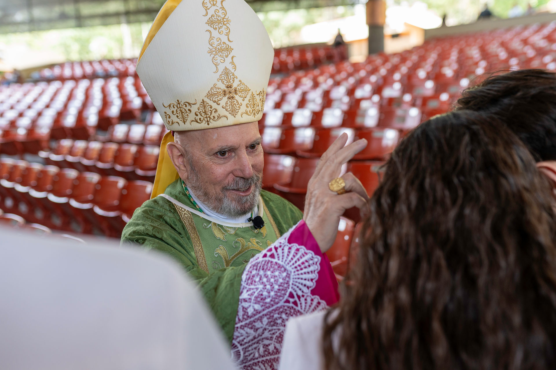 A bishop in green liturgical vestments and a mitre raises his right hand in a blessing gesture. He stands in front of rows of empty red seats in an indoor or covered venue.