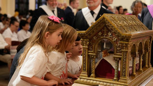 Three young girls in white shirts gaze at an ornate gold reliquary during a solemn church ceremony, with clergy blurred behind them.