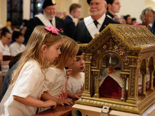 Three young girls in white shirts gaze at an ornate gold reliquary during a solemn church ceremony, with clergy blurred behind them.