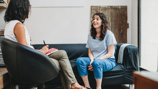 Two women in therapy session; one on chair taking notes, the other on a couch talking, smiling. Neutral-toned room with light walls.
