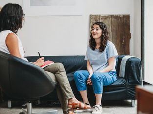 Two women in therapy session; one on chair taking notes, the other on a couch talking, smiling. Neutral-toned room with light walls.