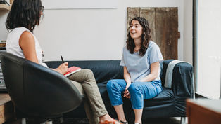 Two women in therapy session; one on chair taking notes, the other on a couch talking, smiling. Neutral-toned room with light walls.