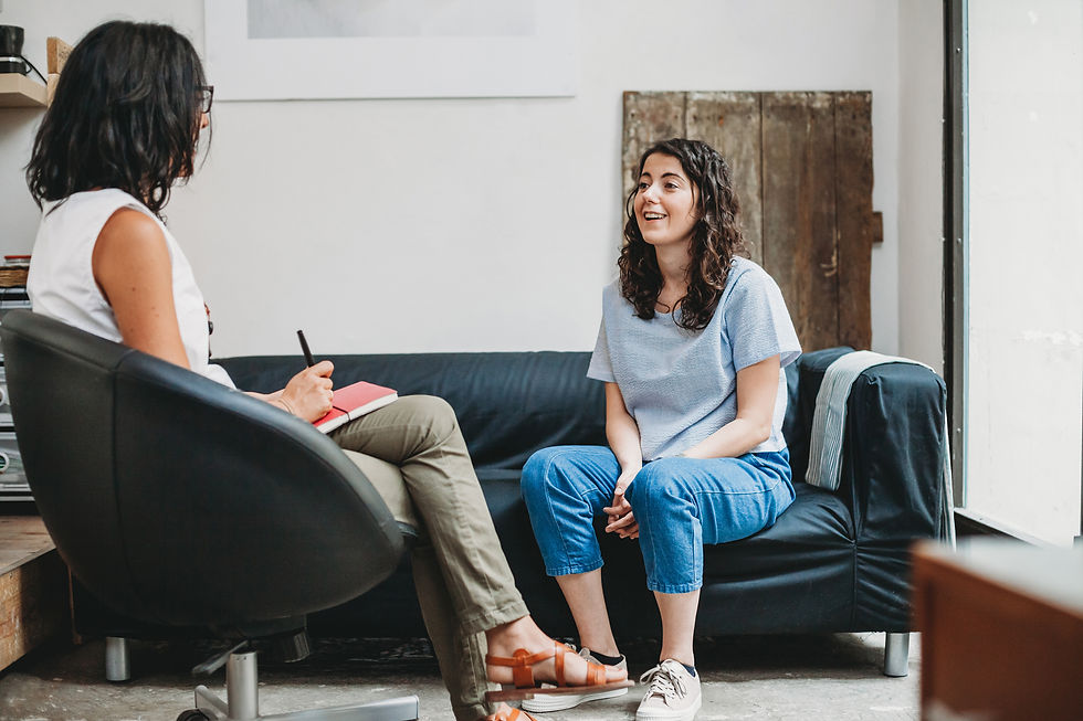 Two women in therapy session; one on chair taking notes, the other on a couch talking, smiling. Neutral-toned room with light walls.