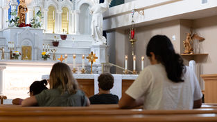 People praying in a church with a white altar, candles, and angel statues. The atmosphere is calm and reverent.