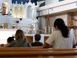 People praying in a church with a white altar, candles, and angel statues. The atmosphere is calm and reverent.