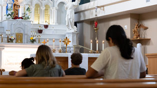 People praying in a church with a white altar, candles, and angel statues. The atmosphere is calm and reverent.