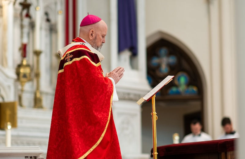 Bishop in red robe and pink zucchetto prays at a lectern in a church. Background features ornate altar, stained glass, and clergy.