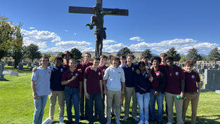 A group of people stands smiling in a cemetery with a large cross behind them. They're on a sunny day with a bright blue sky.