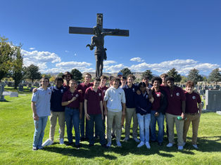 A group of people stands smiling in a cemetery with a large cross behind them. They're on a sunny day with a bright blue sky.
