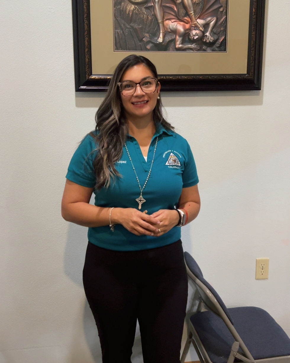 Smiling woman in glasses, teal shirt, standing next to a chair. Background features a framed artwork depicting a dramatic scene.