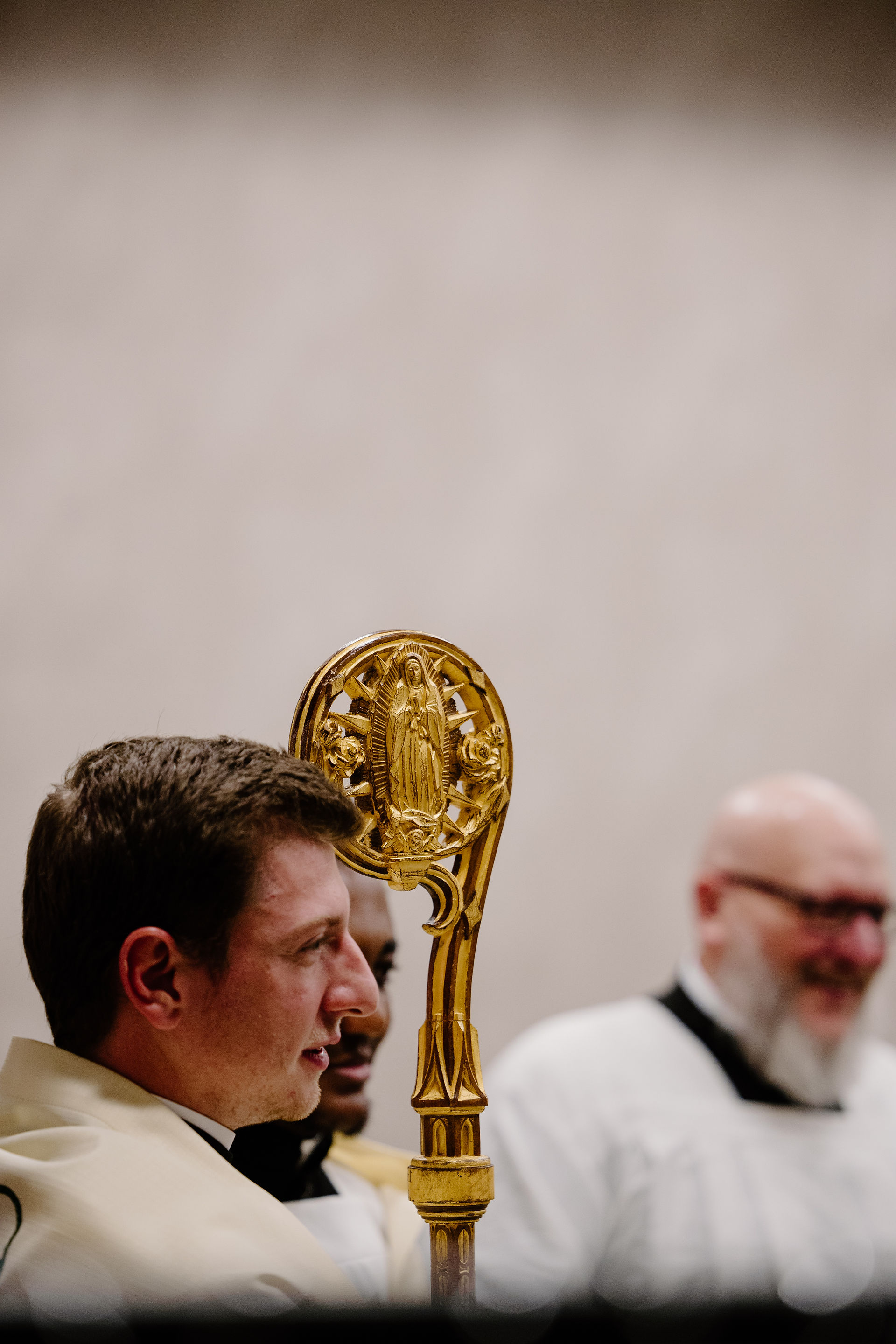 Two men in formal attire, one holding an ornate gold staff with a religious figure. Neutral background, serious and focused expressions.