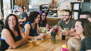 Group of people smiling and chatting around a table with drinks and a vase of flowers in a cozy café setting.