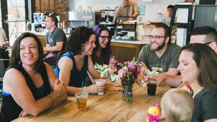 Group of people smiling and chatting around a table with drinks and a vase of flowers in a cozy café setting.