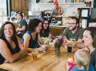Group of people smiling and chatting around a table with drinks and a vase of flowers in a cozy café setting.