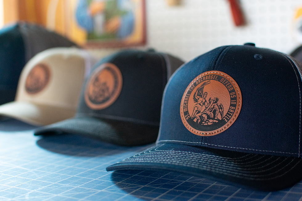 Three caps with leather patches sit on a blue grid surface. The patches display "St. Michael the Archangel, Defend Us in Battle."