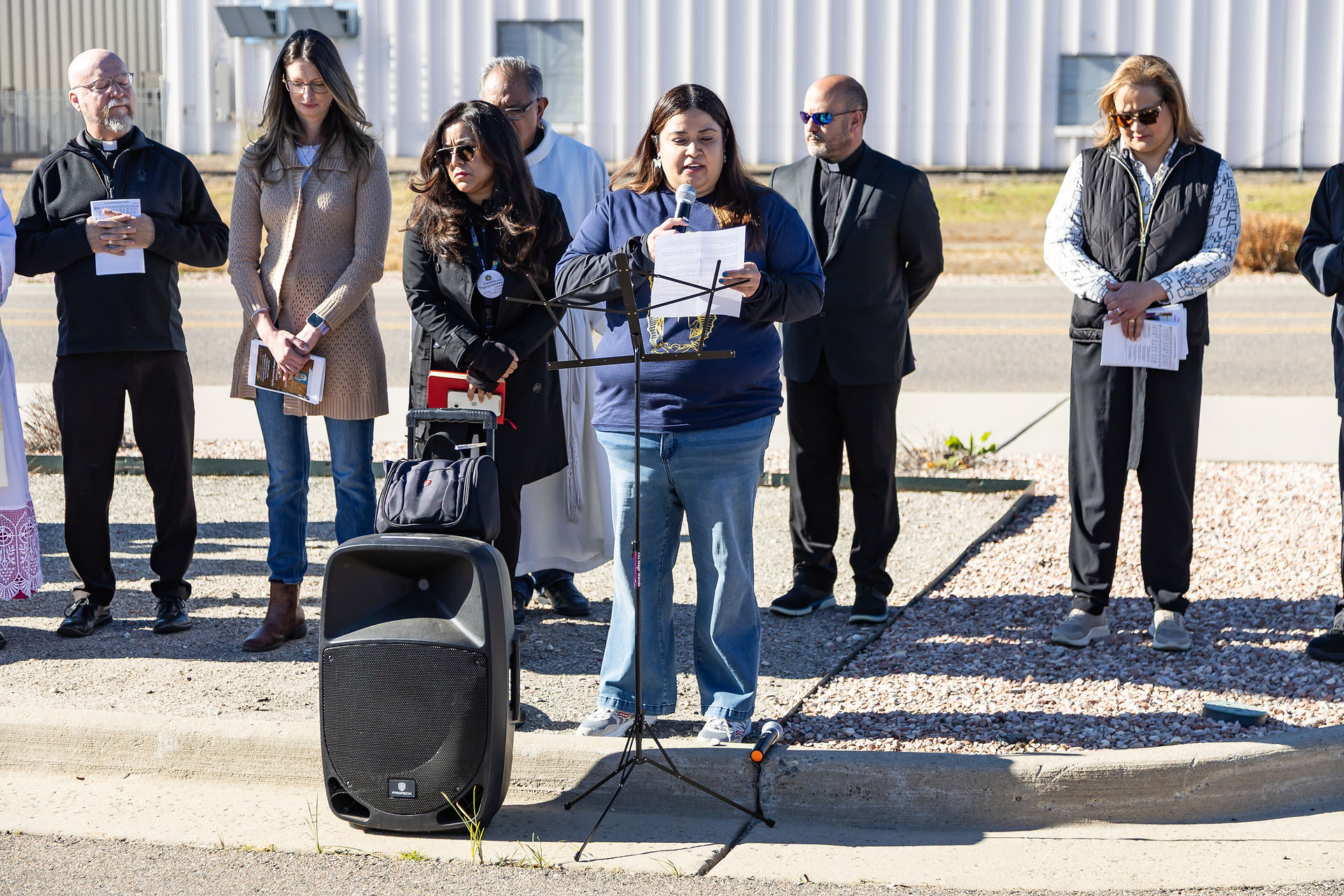 A group of people stands outdoors on gravel. A woman speaks into a microphone, holding papers. Amplifier and music stand visible in front.