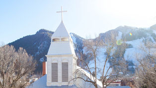 White church with a cross atop, in front of snow-capped mountains and bare trees, under a clear blue sky. Bright and serene atmosphere.