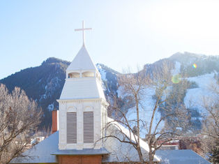 White church with a cross atop, in front of snow-capped mountains and bare trees, under a clear blue sky. Bright and serene atmosphere.
