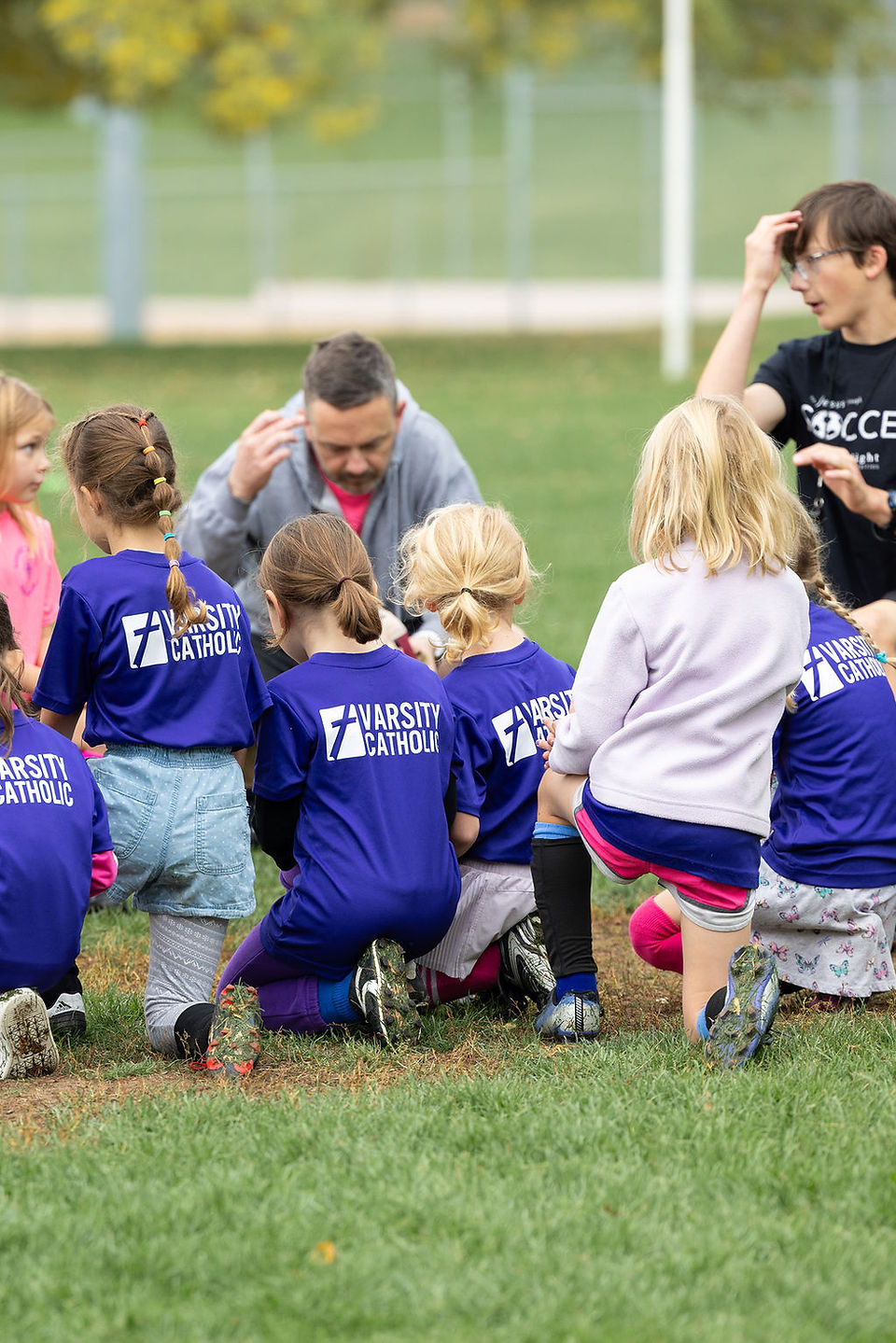 Children in purple Varsity Catholic shirts kneel with a coach on a grassy field during an outdoor team huddle.