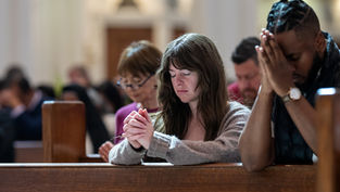 People praying in a church, eyes closed, hands clasped. Wooden pews and soft lighting create a serene atmosphere.