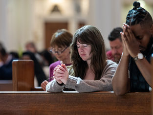 People praying in a church, eyes closed, hands clasped. Wooden pews and soft lighting create a serene atmosphere.