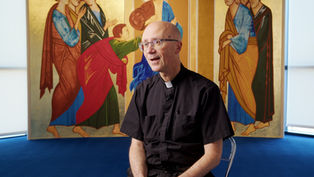 A person in clergy attire sits in front of a vibrant, detailed religious mural featuring figures in colorful robes. Bright, calm setting.