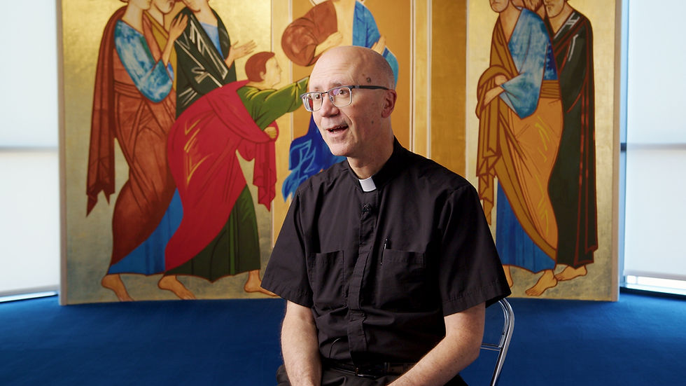 A person in clergy attire sits in front of a vibrant, detailed religious mural featuring figures in colorful robes. Bright, calm setting.