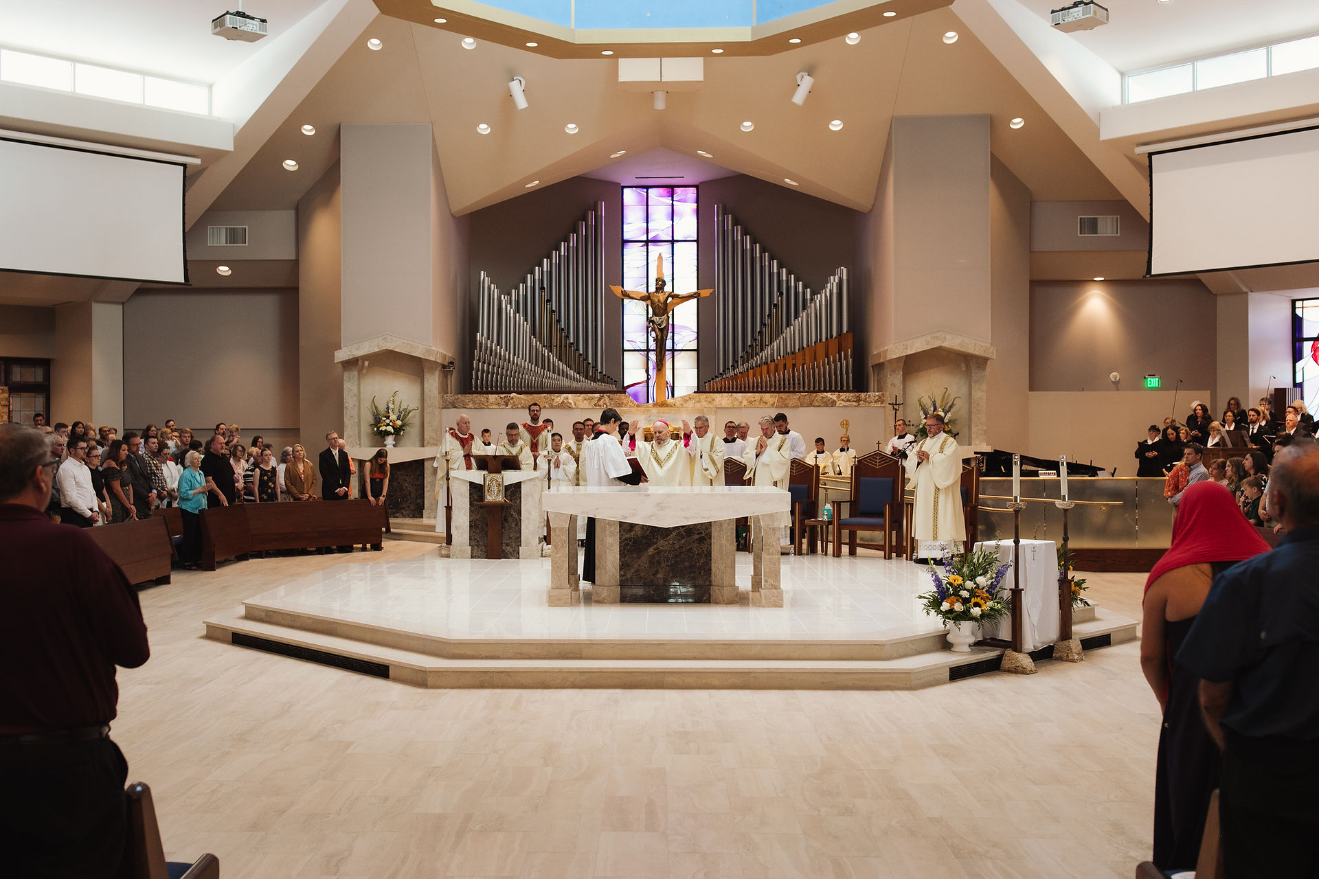Priests in white robes perform a ceremony at a church altar, with a large organ and stained-glass window behind. Congregation watches silently.