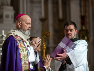 Clergyman in ornate purple and gold vestments prays beside another man holding a book in a church, solemn atmosphere, detailed carvings.