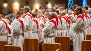 Clergy in white robes with red stoles stand and pray in a church setting, surrounded by wooden pews and stained glass. Peaceful atmosphere.