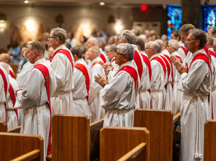 Clergy in white robes with red stoles stand and pray in a church setting, surrounded by wooden pews and stained glass. Peaceful atmosphere.