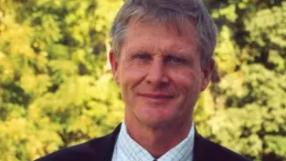 A man in a suit stands outdoors with a background of lush green trees. He is smiling slightly, creating a calm and relaxed mood.