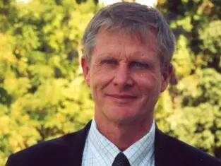 A man in a suit stands outdoors with a background of lush green trees. He is smiling slightly, creating a calm and relaxed mood.