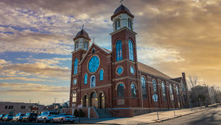 Brick church with twin towers and blue stained-glass windows at sunset, parked cars along the street, under a warm cloudy sky.