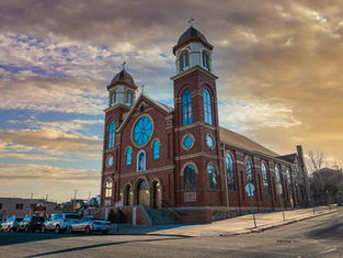 Brick church with twin towers and blue stained-glass windows at sunset, parked cars along the street, under a warm cloudy sky.