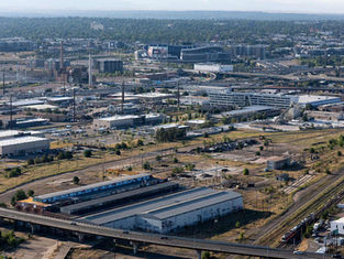 Aerial view of an industrial area with warehouses, rail lines, and scattered greenery. A distant stadium is visible under a clear sky.
