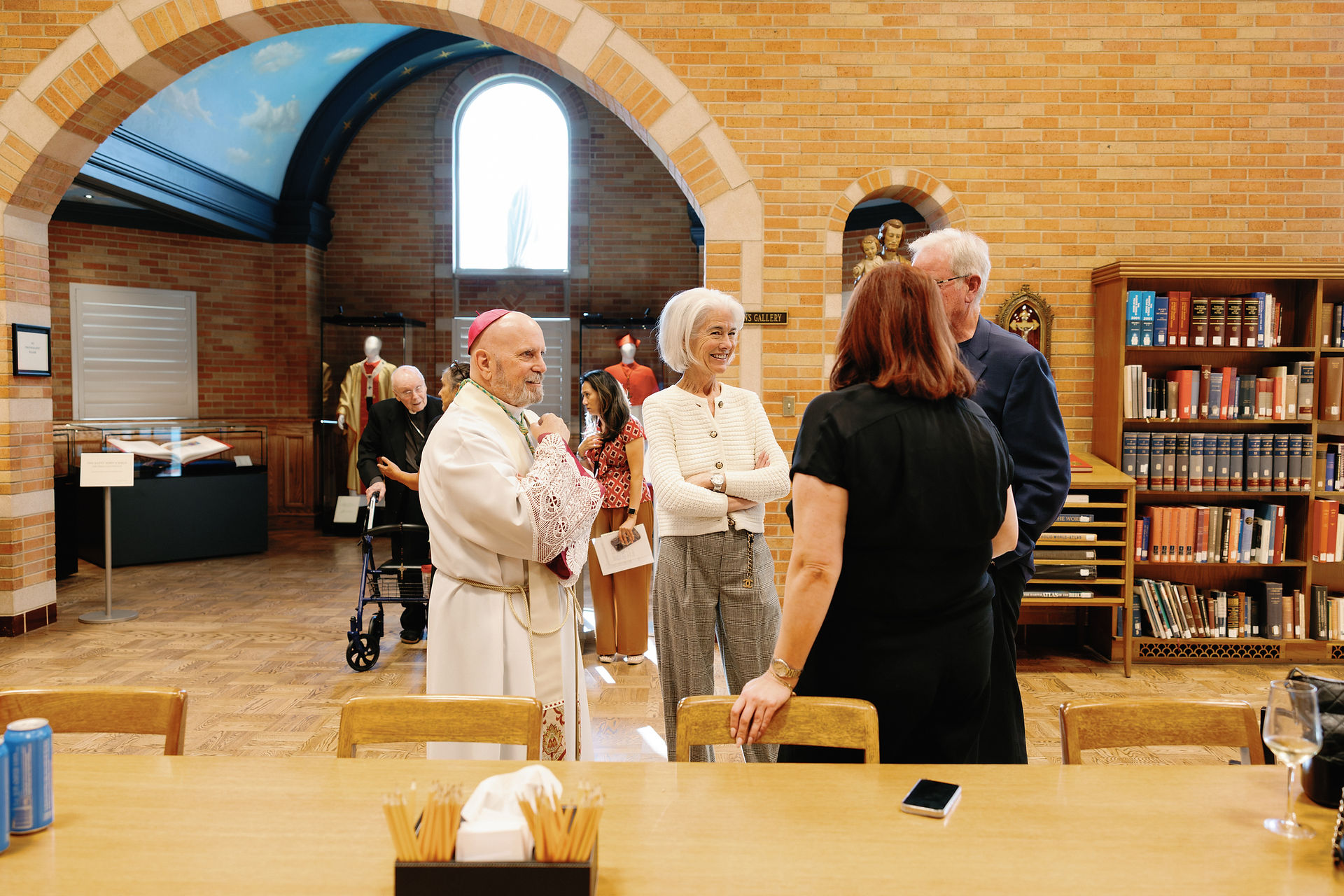 People conversing in a library with brick walls, arched windows, and bookshelves. A clergyman in a white robe is present. A sky-painted ceiling adds color.