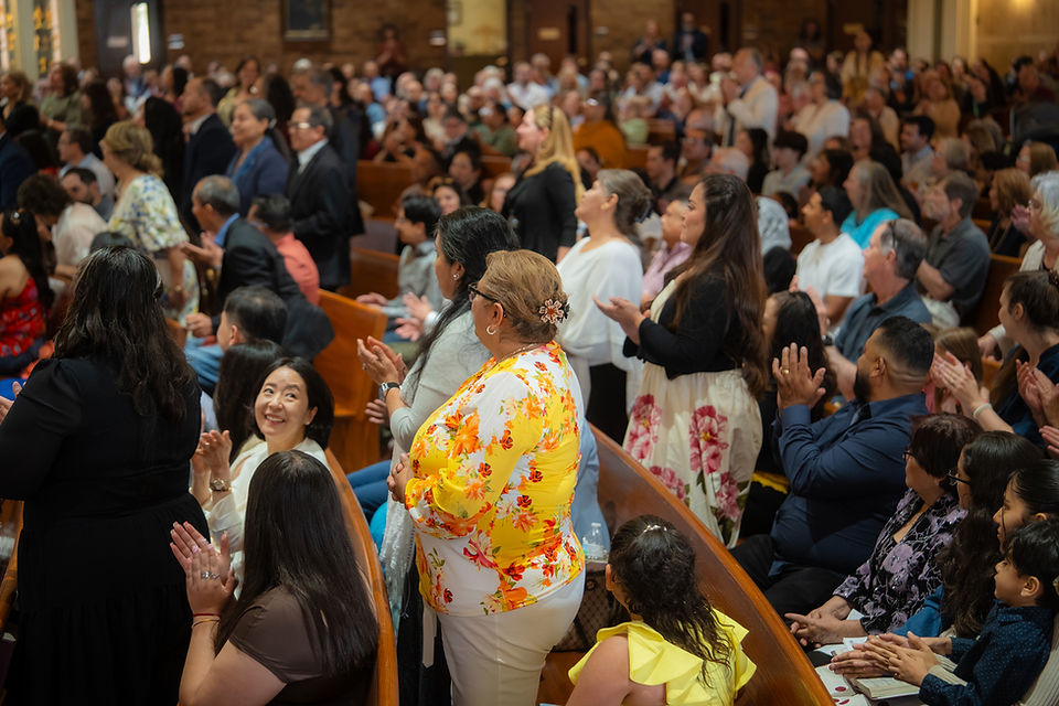 Congregation in a church stands and claps in wooden pews, including a woman in a bright floral blouse, joyful crowd scene