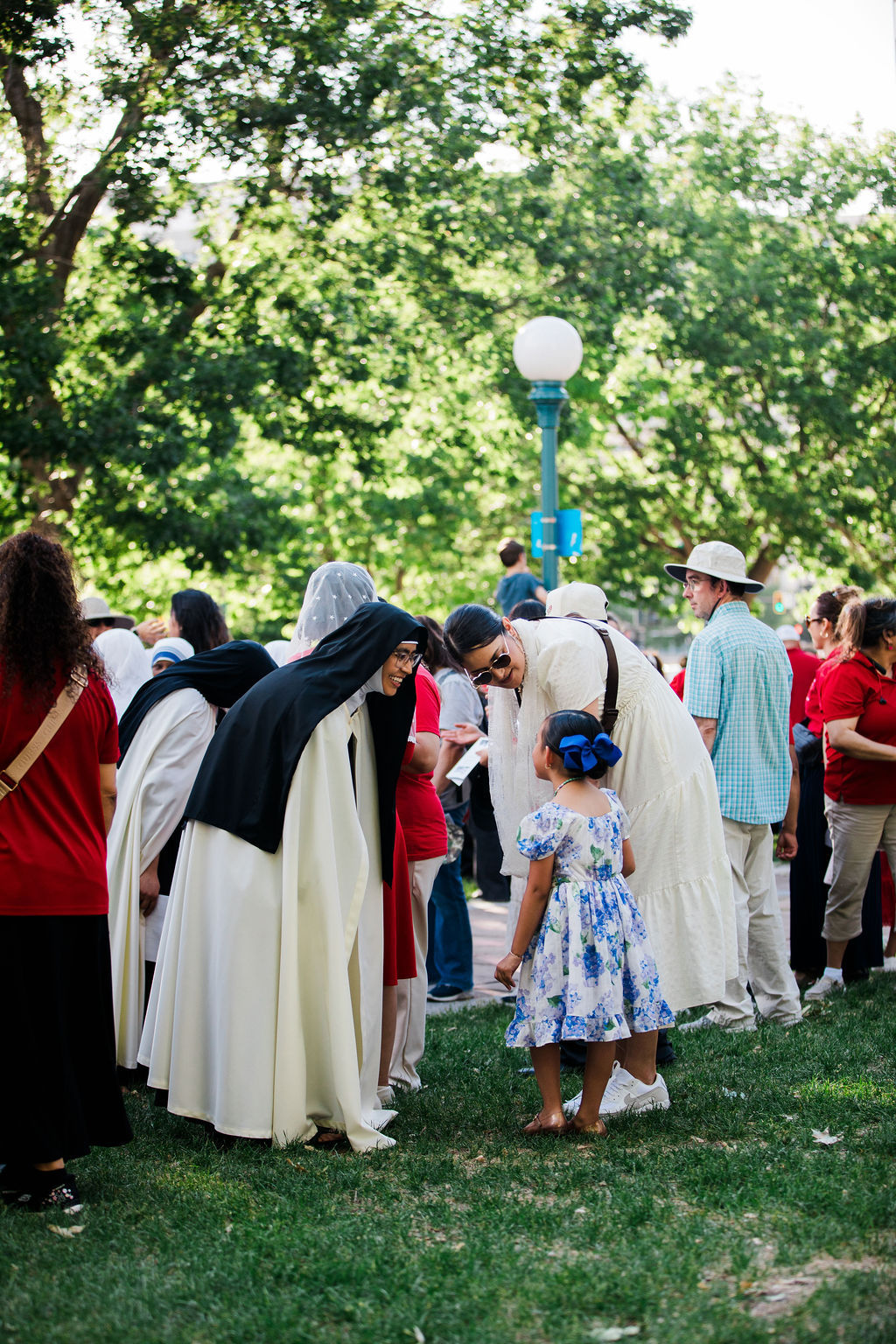 Nuns in white habits smile and greet a little girl in a blue floral dress at a sunny park gathering.